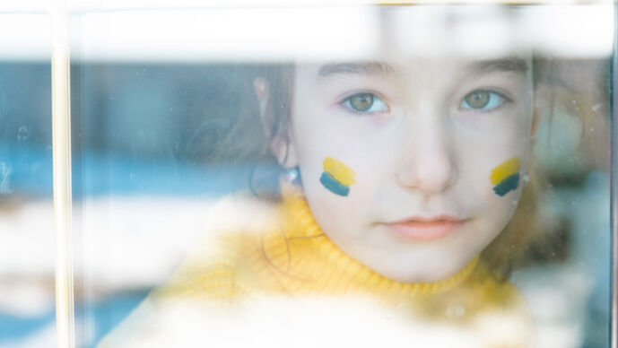 Ledset barn med en ukrainska flagga målad på kinderna kikar ut genom fönster